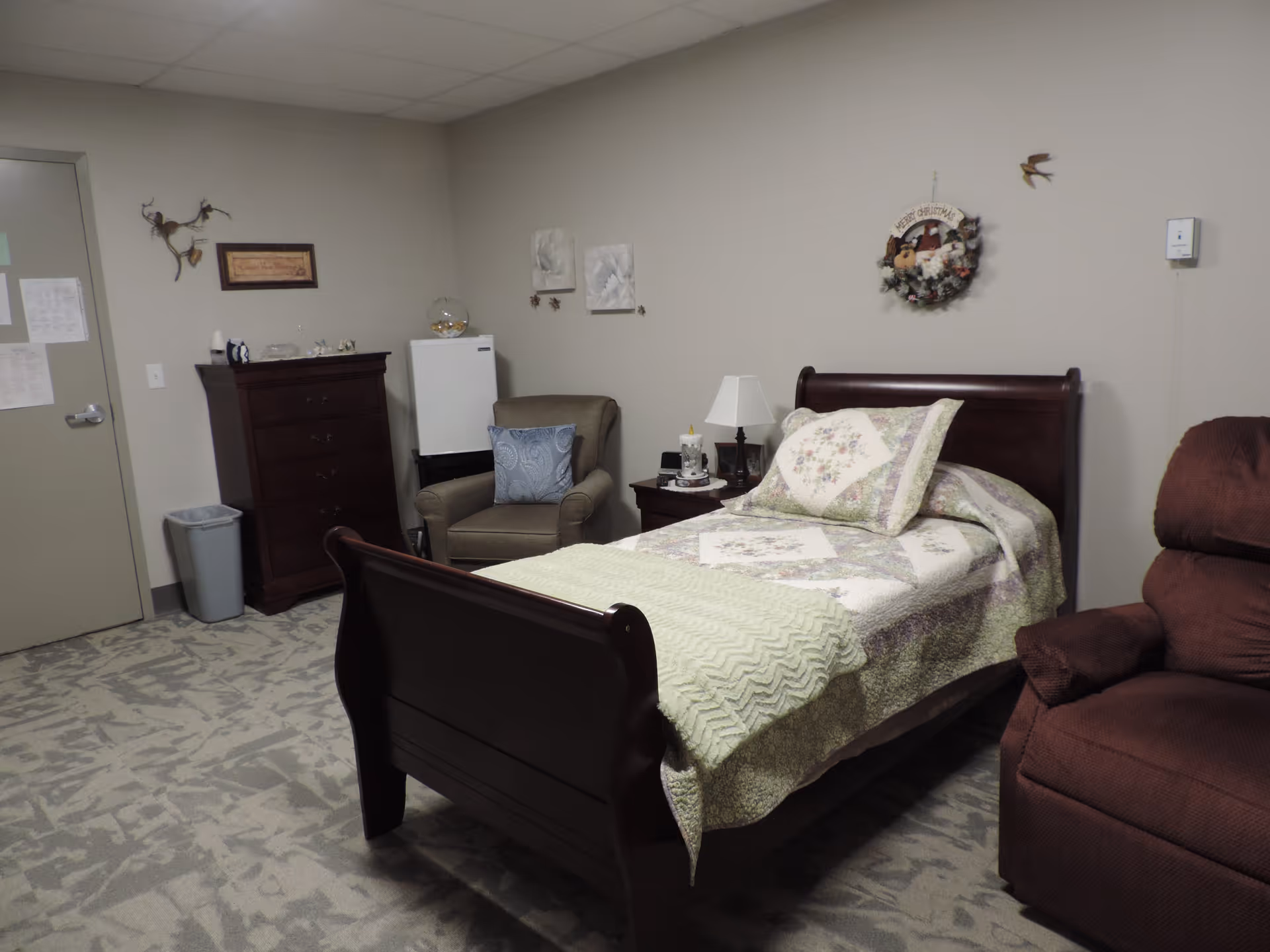 A cozy assisted living bedroom with a wooden bed covered in a floral quilt and a green blanket. Next to the bed is a nightstand with a lamp and a cup. There is a brown recliner chair on the right and a beige armchair with a blue pillow near a small white refrigerator. A tall wooden dresser stands against the wall near the door. The walls are decorated with a wreath, two framed pictures, and a small bird decoration.
