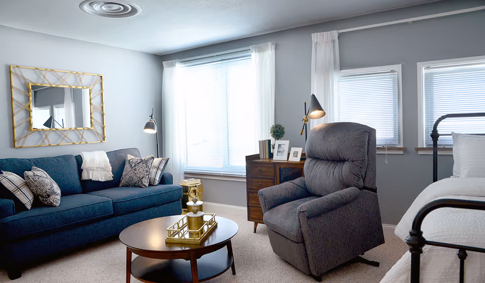 Sunlit living area with a blue sofa, gray recliner, wooden coffee table, dresser and a bed beside large windows.