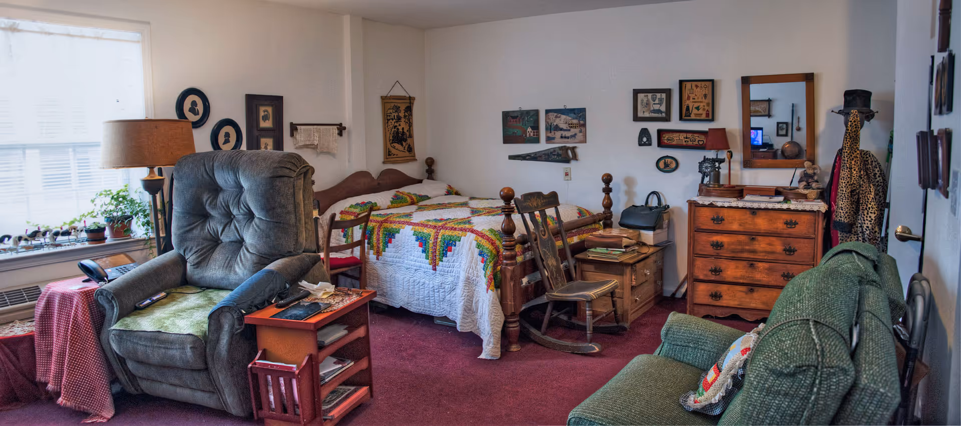 A cozy, vintage-style assisted living room featuring a wooden bed with a colorful quilt, a green recliner chair, a wooden rocking chair, a green upholstered sofa, a wooden dresser with a mirror, and various framed pictures and decorations on the walls. There is a window with blinds letting in natural light, a floor lamp, and a small side table with books and a phone.