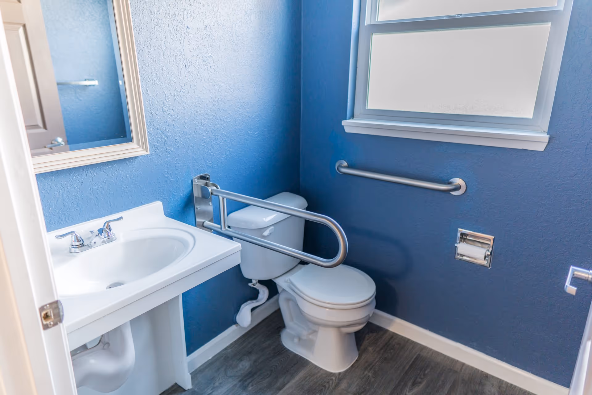 Small accessible bathroom with blue walls, a toilet equipped with grab bars, a sink and a window.
