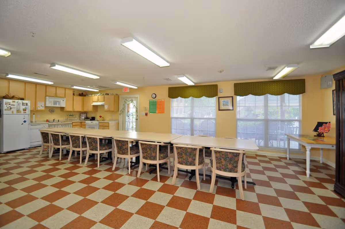Communal dining/activity room with a long row of tables and chairs and a kitchenette in the background.