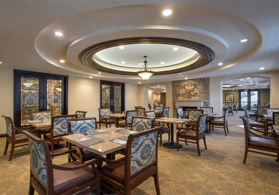 Spacious, elegantly furnished dining room with patterned upholstered chairs, multiple tables, a central circular recessed ceiling feature, and a fireplace.