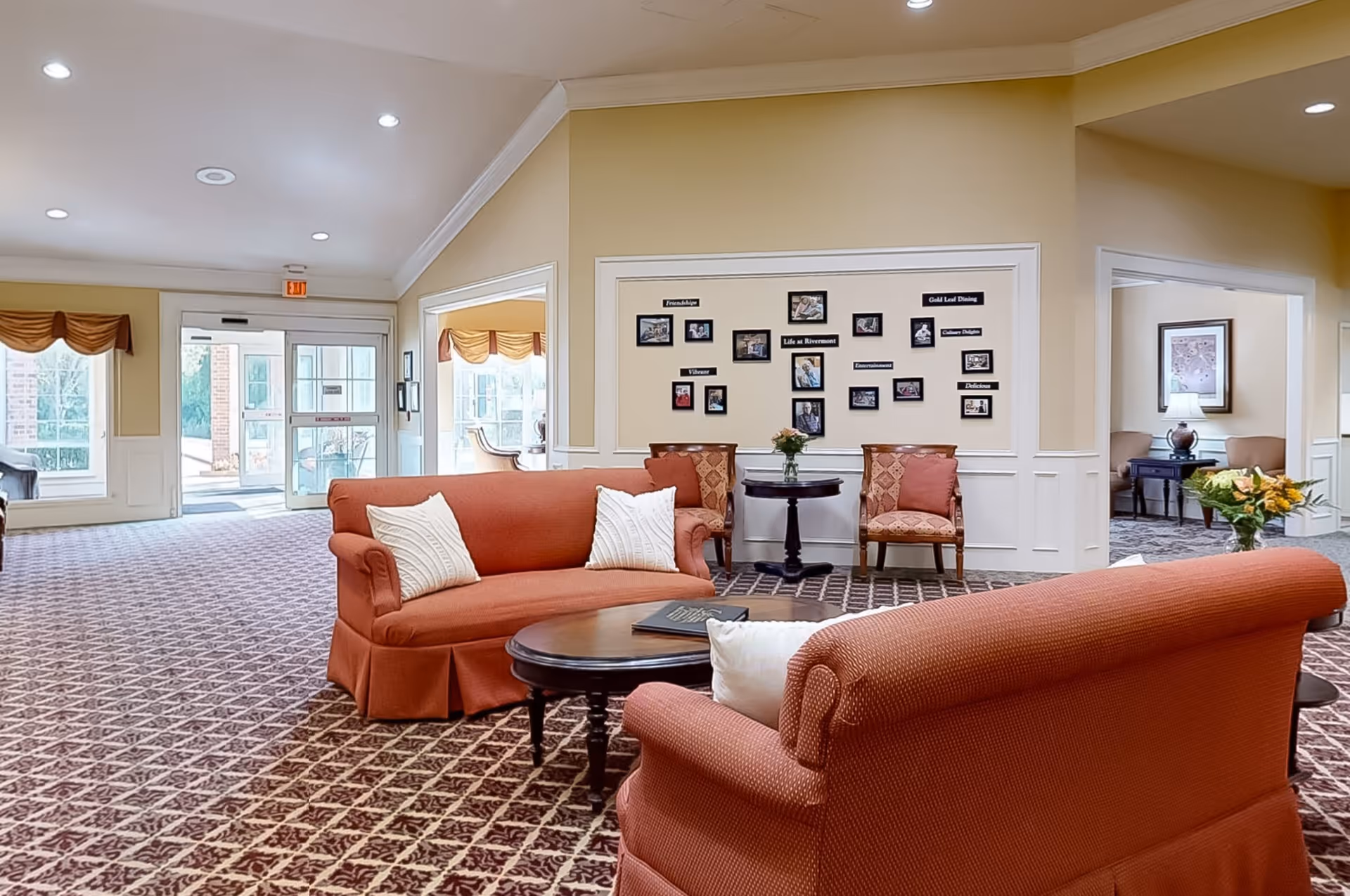 A spacious and well-lit living room area in an assisted living facility with two orange sofas facing a round wooden coffee table. The room has patterned carpet flooring, beige walls with white wainscoting, and several framed photos and labels on the wall. There are two chairs and a small table with a flower vase against the wall, and large windows and glass doors letting in natural light. Another seating area with a lamp and framed artwork is visible through an open doorway.