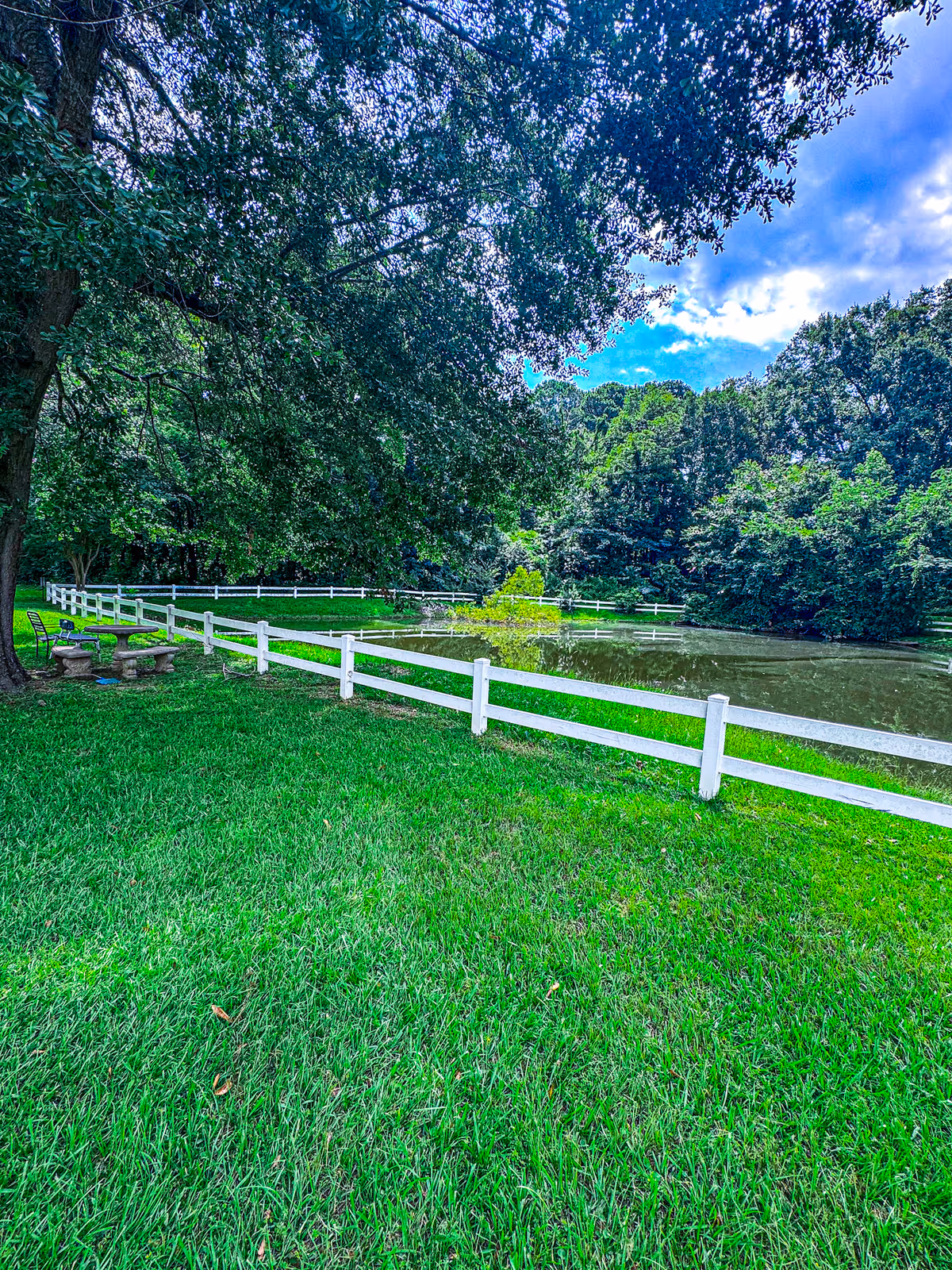 A peaceful outdoor scene featuring a small pond surrounded by a white wooden fence, lush green grass, and large trees providing shade. There is a stone picnic table with benches and a metal chair under the trees near the pond. The sky is partly cloudy with patches of blue visible.