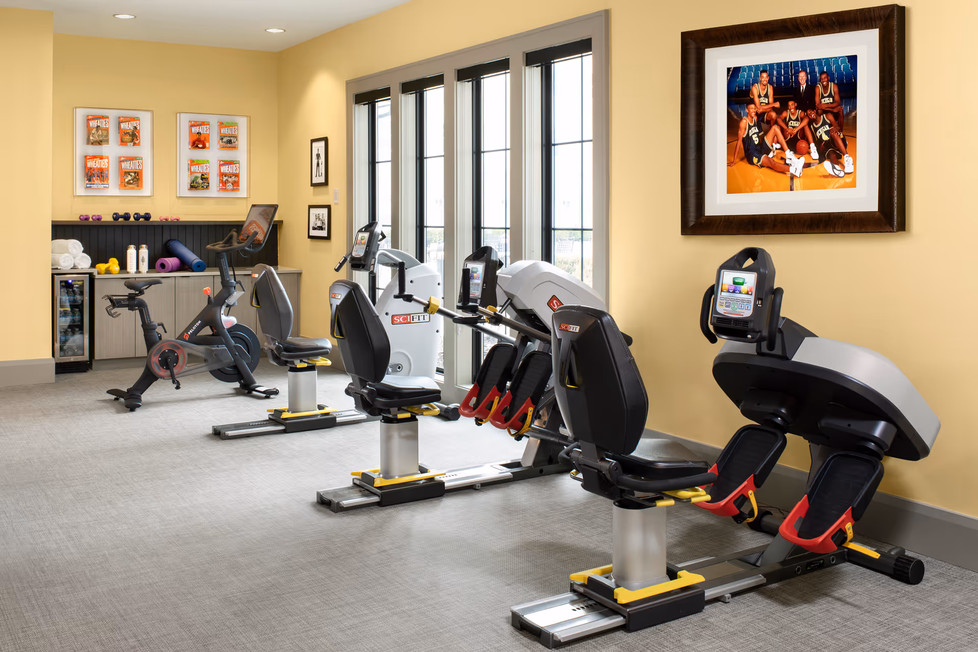 A fitness room with exercise equipment including stationary bikes and rowing machines. The room has yellow walls, large windows letting in natural light, and framed pictures on the walls. There are towels, water bottles, and small weights on a shelf in the corner.