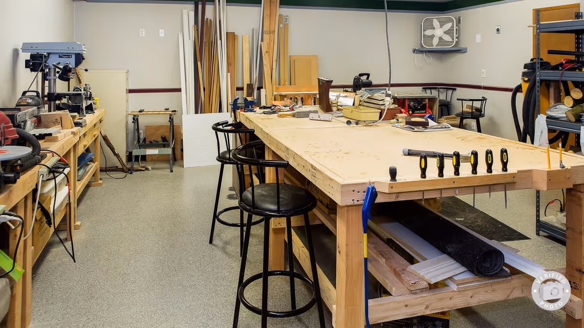 A woodworking workshop with a large wooden workbench in the center, surrounded by stools. Various woodworking tools and materials are placed on the bench and shelves along the walls. The room has a clean, organized appearance with a fan mounted on the wall.
