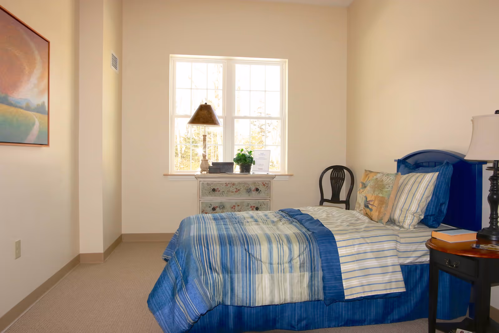 A cozy bedroom with a single bed dressed in blue and white striped bedding. There is a small wooden nightstand with a lamp and books on the right side of the bed. A decorative chest of drawers with a lamp, a plant, and a framed item sits under a window letting in natural light. A black wooden chair is placed against the wall near the window. A colorful painting hangs on the left wall.