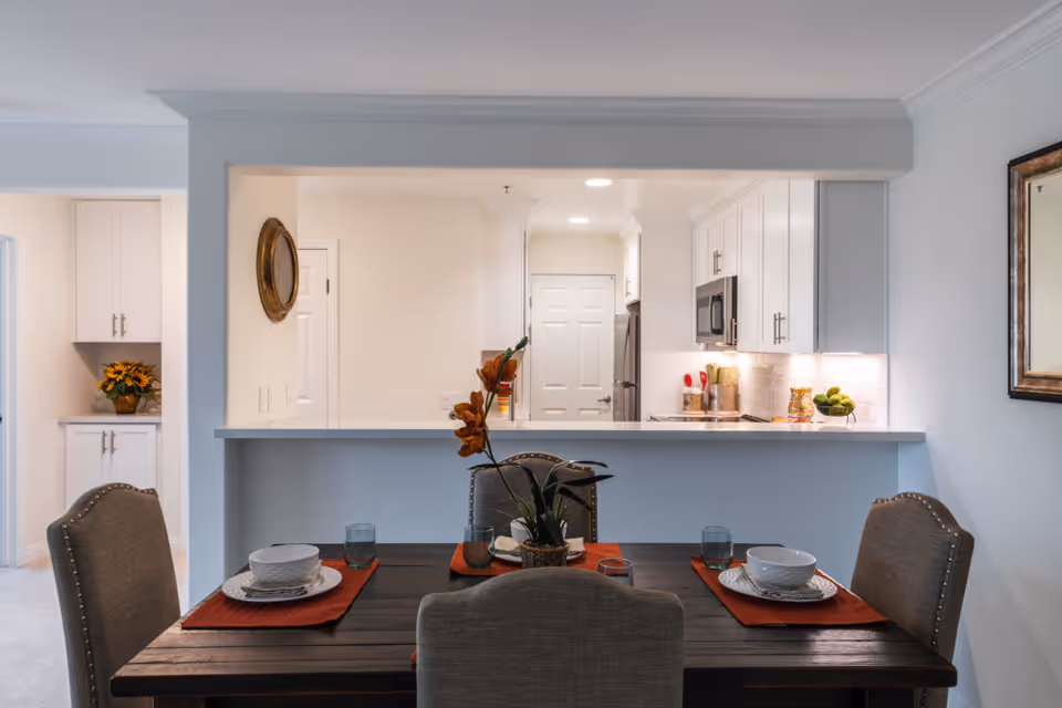 View of a dining area with a dark wooden table set for four with bowls, plates, glasses, and orange placemats. Behind the dining area is a kitchen with white cabinets, a microwave, and a refrigerator. The kitchen counter has decorative items including a bowl of green apples and a container with utensils. A mirror hangs on the wall to the right, and a small cabinet with a flower arrangement is visible to the left.