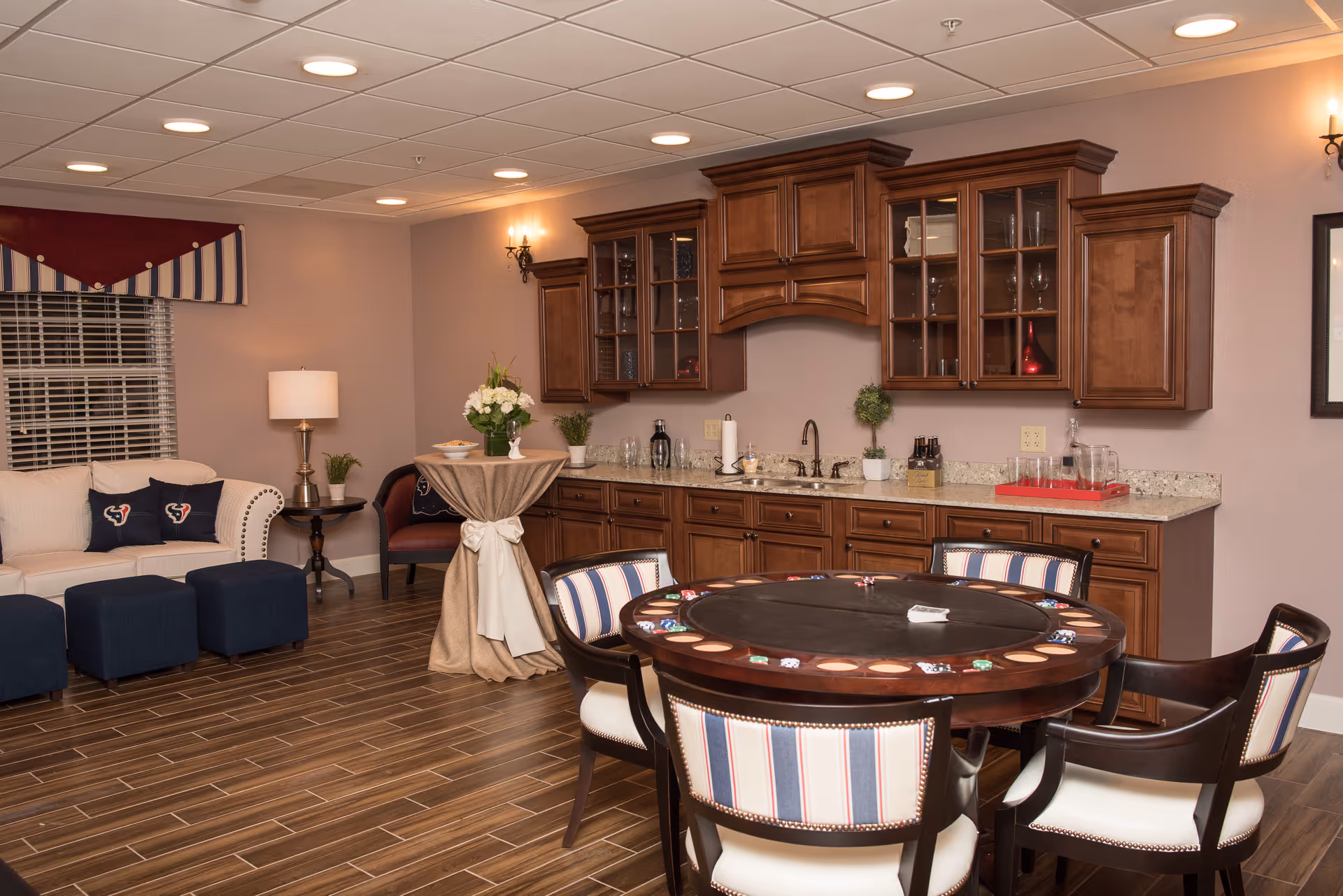 A furnished common room with a round poker table and striped chairs, a sofa seating area, and a kitchenette with wooden cabinets.