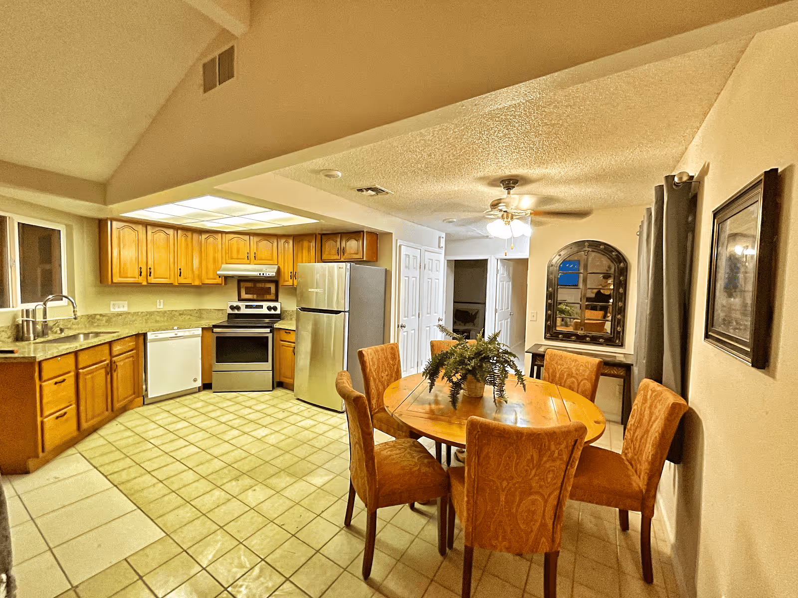 Open-plan kitchen with wooden cabinets, stainless steel appliances and a round dining table with six upholstered chairs.