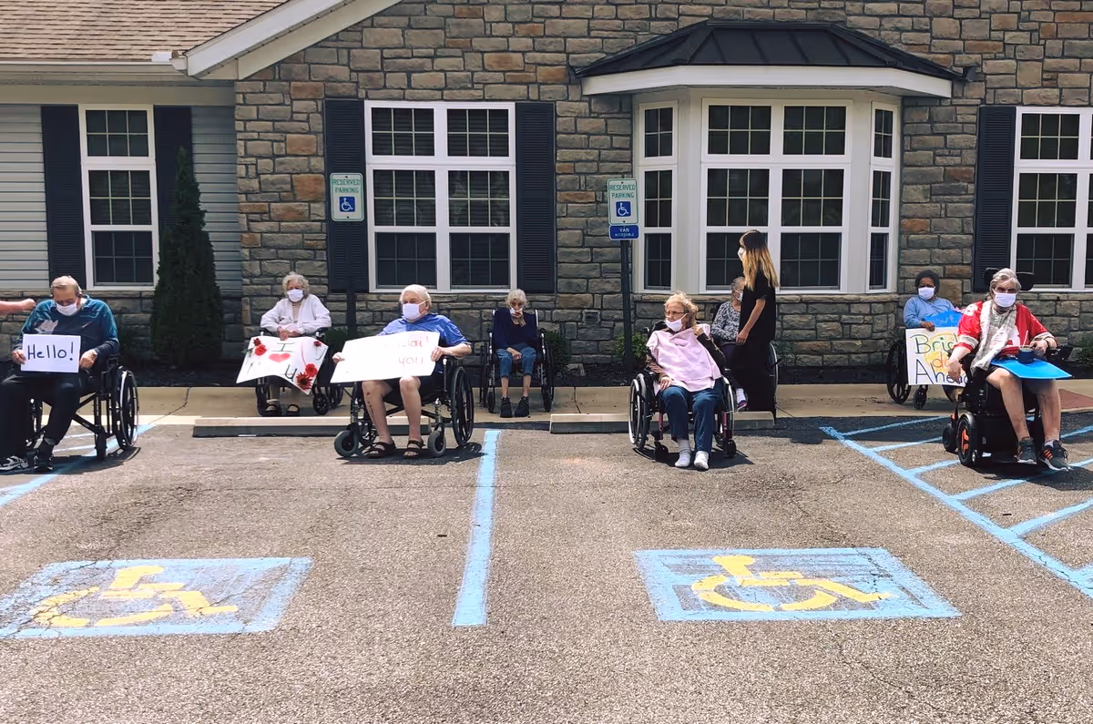 Several people in wheelchairs and a caregiver wearing masks sit outside the front of The Laurels of Athens holding signs in accessible parking spaces.