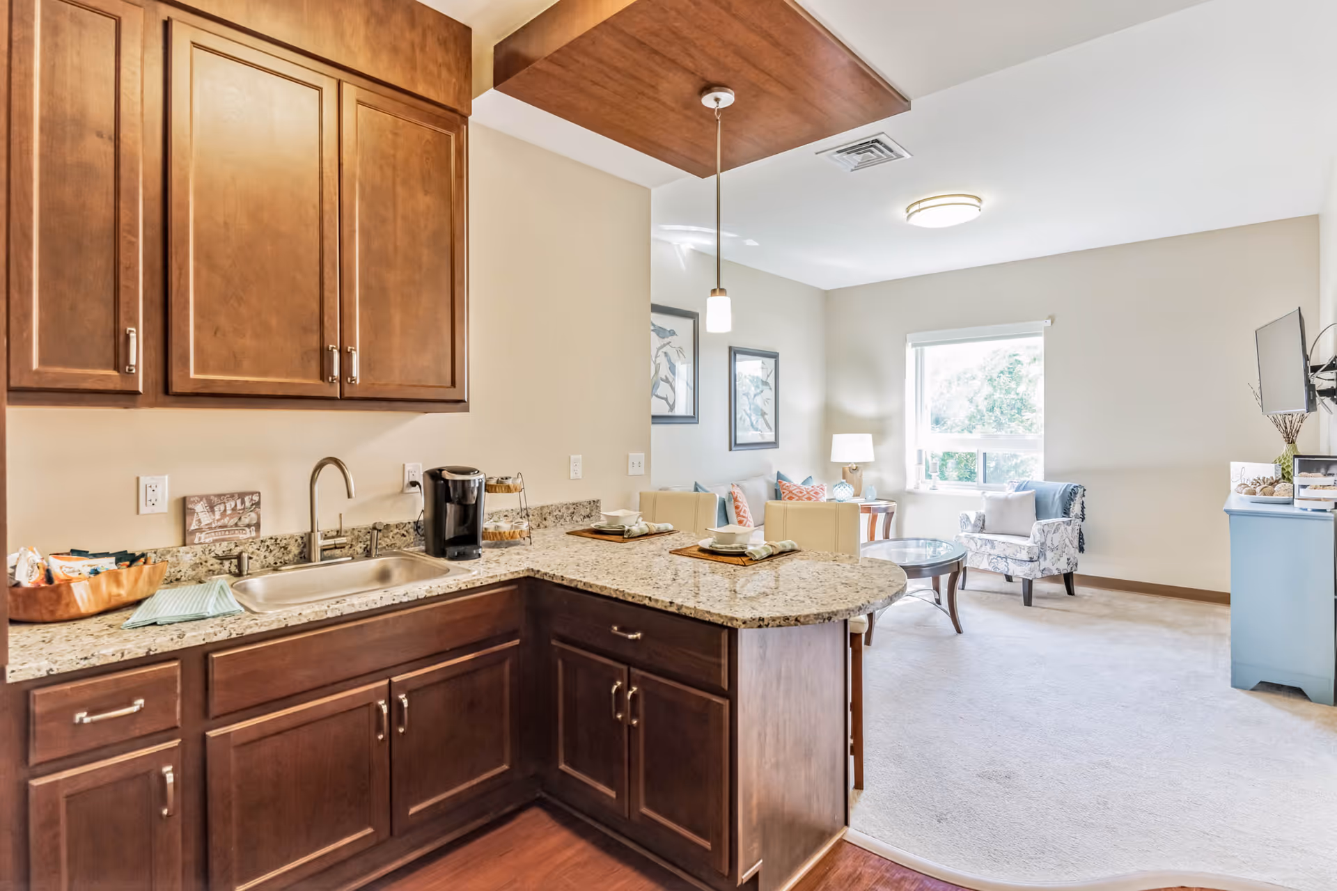 Open-plan kitchen with dark wood cabinets, granite countertops and a small living area visible in the background.