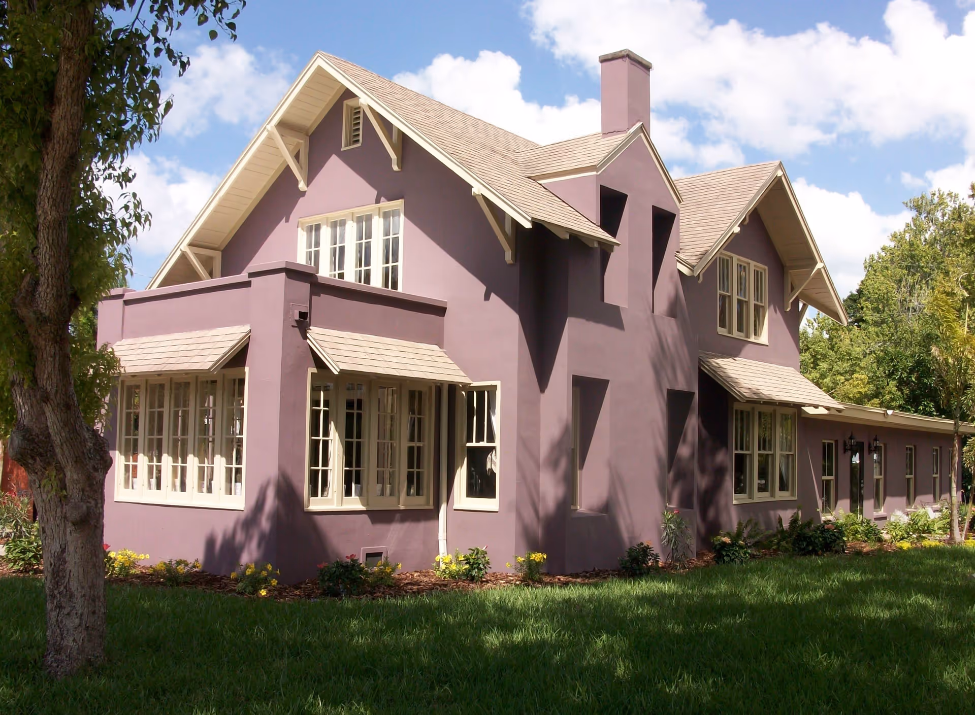 Exterior view of a two-story purple building with multiple windows, a sloped roof, and a chimney, surrounded by green grass, trees, and some small plants under a partly cloudy sky.