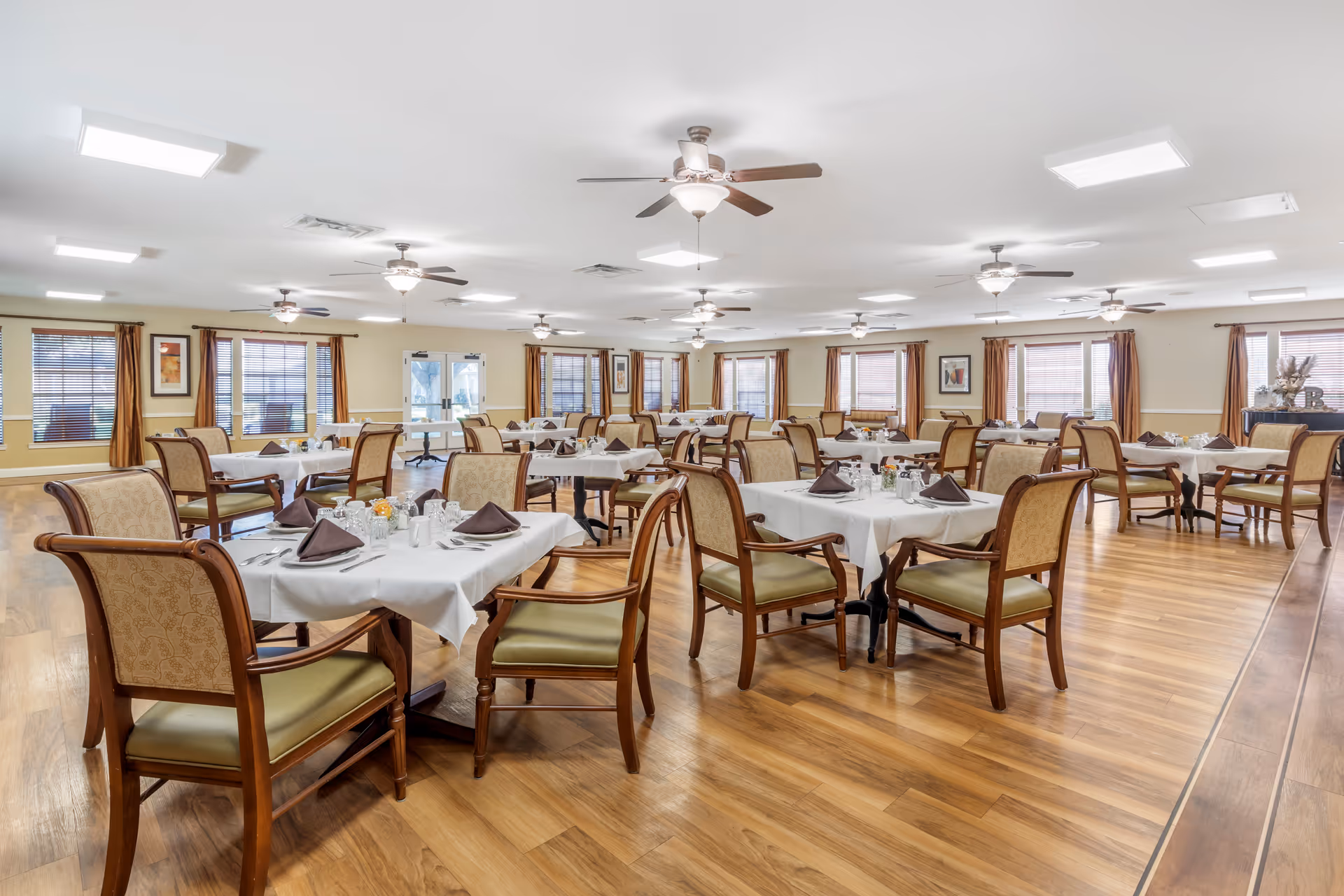 A spacious dining room with multiple tables covered in white tablecloths, each set with folded brown napkins, silverware, and small condiment containers. The room has wooden floors, beige walls, several windows with brown curtains, ceiling fans, and bright overhead lighting.