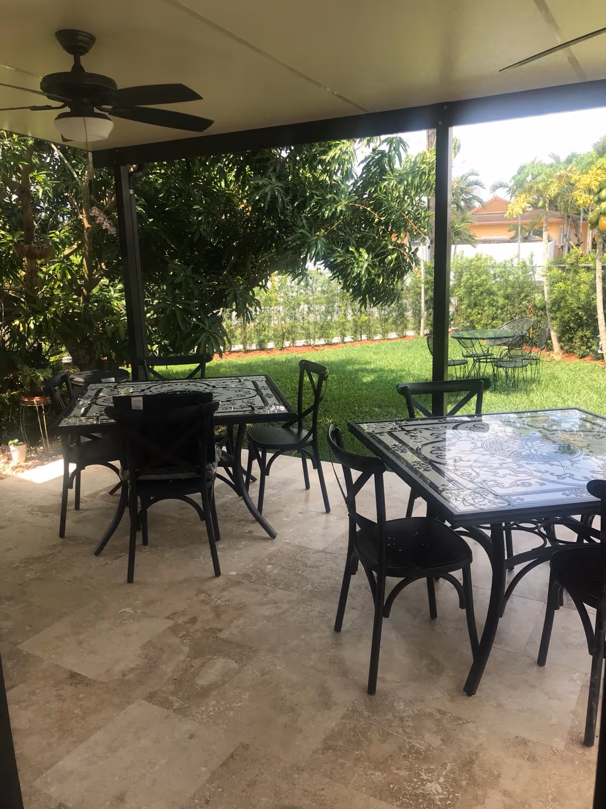 Covered outdoor patio area with two black metal tables featuring decorative glass tops and several black chairs. Ceiling fan is mounted above. The patio overlooks a green lawn with trees and shrubs, and additional metal table and chairs are visible on the grass.