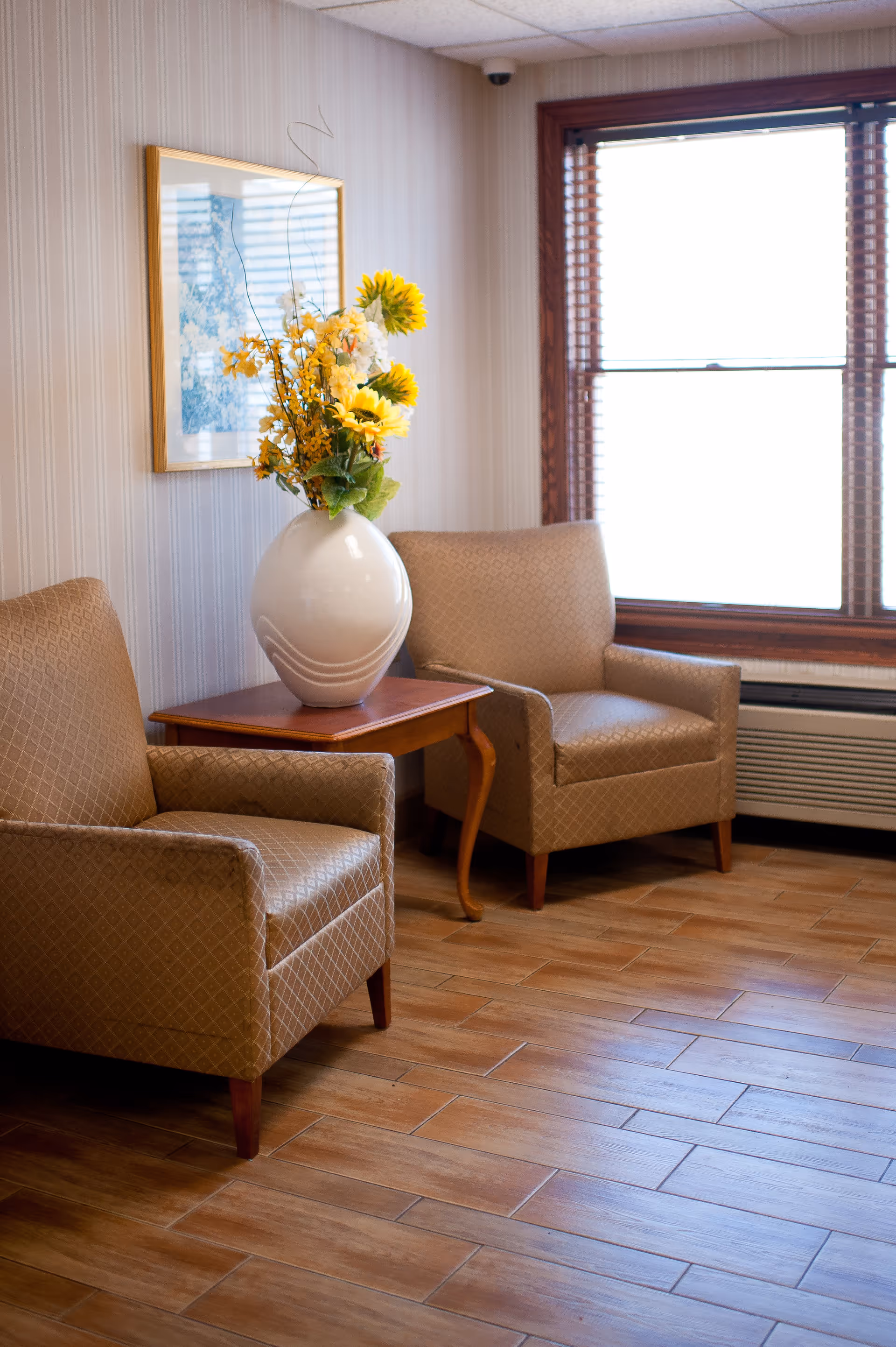 A cozy sitting area with two beige patterned armchairs placed on a wooden floor. Between the chairs is a wooden side table holding a large white vase filled with yellow and white flowers. Behind the chairs is a window with wooden blinds and a framed artwork on the wall.