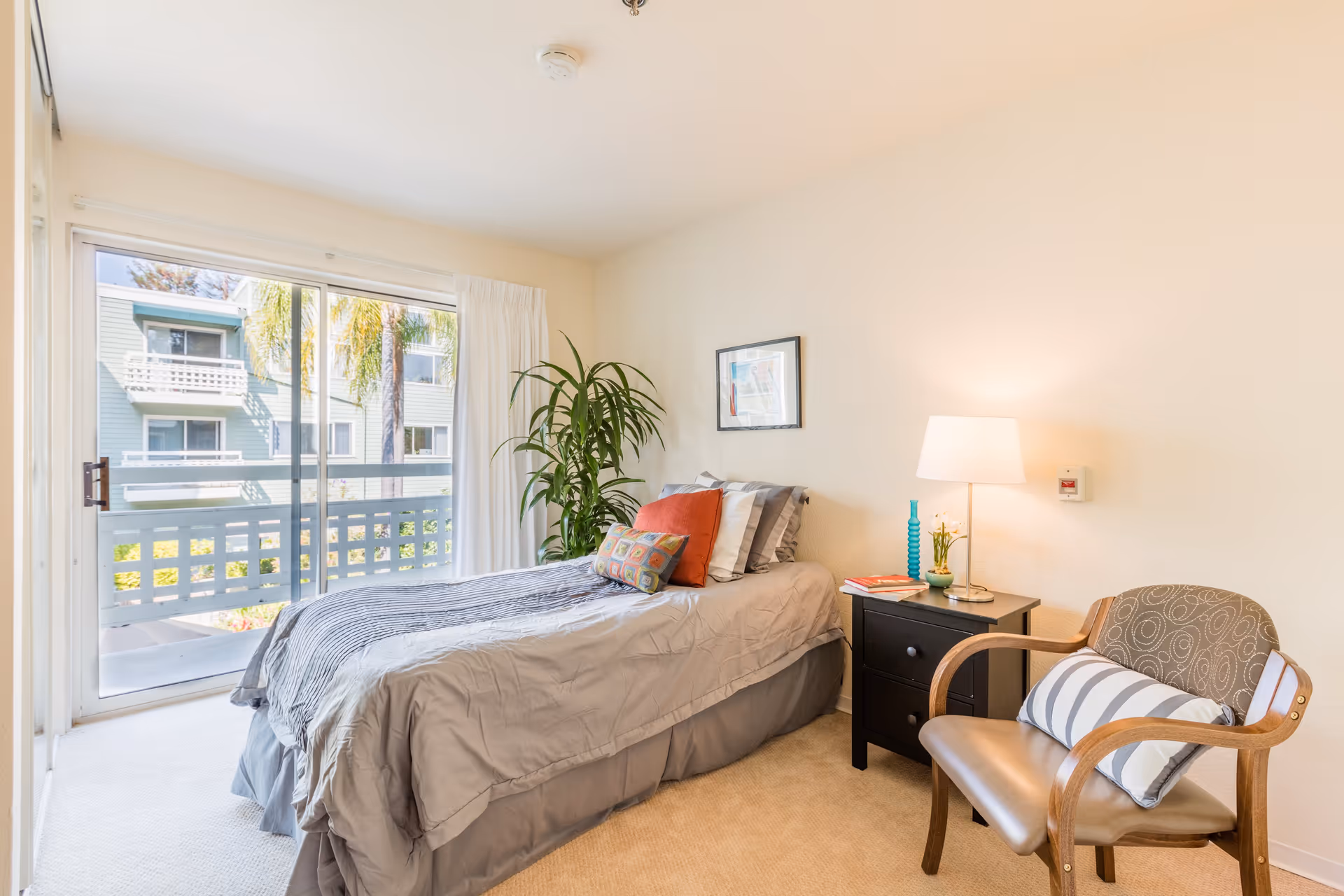 A bright bedroom with a single bed made with gray bedding and colorful pillows. Next to the bed is a black nightstand with a lamp, a small plant, and some books. A wooden chair with a striped cushion is placed beside the nightstand. A large sliding glass door with white curtains opens to a balcony, showing an exterior view of a building and palm trees. A tall green plant is positioned near the bed, and a framed picture hangs on the wall above the bed.