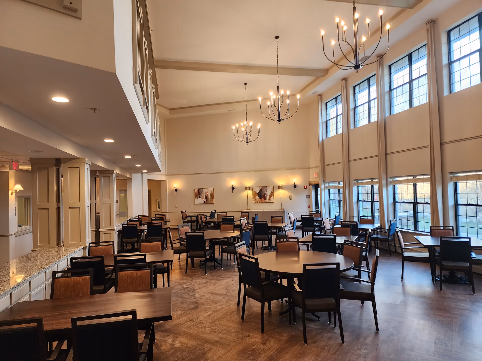 Spacious dining room with multiple tables and chairs arranged neatly. Large windows on the right side allow natural light to fill the room. The ceiling is high with three modern chandeliers hanging down. The walls are decorated with two abstract paintings and several wall-mounted lights.