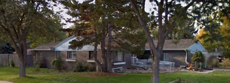 Exterior view of a single-story brick building with a shingled roof, surrounded by trees and green lawn. There is a small porch with a white railing and a ramp leading to the entrance.