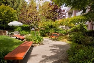 A peaceful outdoor garden area with a paved walkway, wooden benches, green shrubs, trees, and a white umbrella shading a table with chairs. Part of a building is visible on the right side.