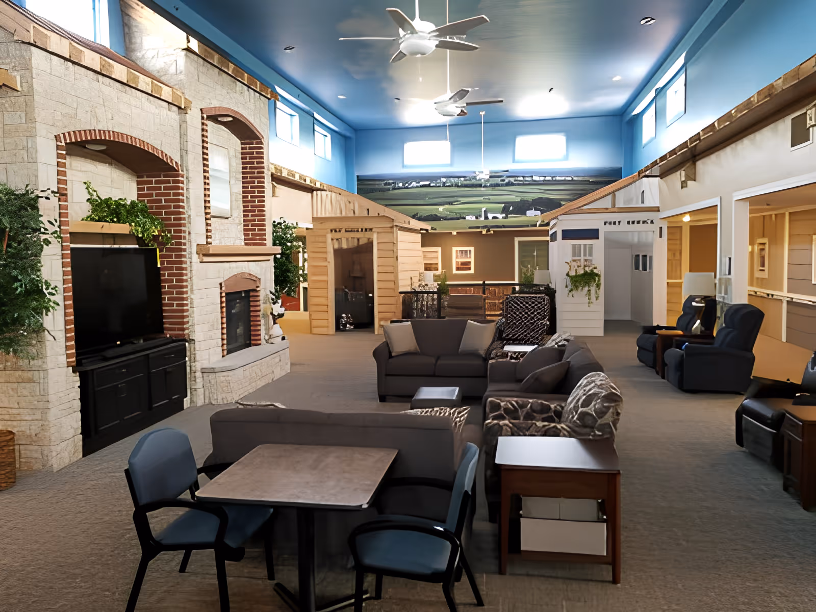 Spacious indoor common area with sofas, recliners, a TV and fireplace beneath a blue ceiling and small model house facades.