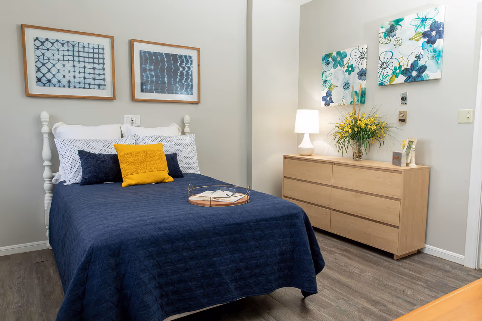 A neatly made bed with a navy blue quilt, white and navy pillows, and a yellow accent pillow in a bedroom. Above the bed are two framed abstract artworks. To the right is a light wood dresser with a white lamp, a vase of yellow flowers, and two floral paintings hanging above it. The room has light-colored walls and wood flooring.