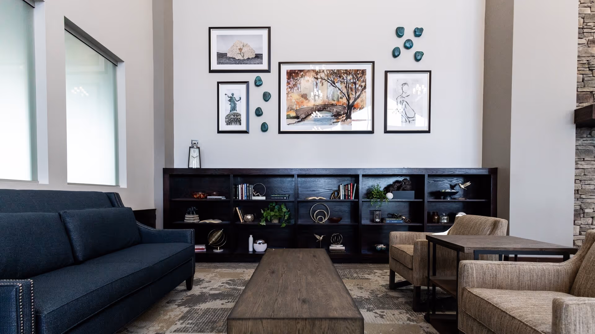 A modern lounge with a dark sofa and armchairs around a long wooden coffee table, a low bookshelf and framed artwork on the wall.