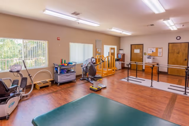 A spacious rehabilitation room with wooden flooring, featuring exercise equipment including a recumbent bike, parallel bars for walking practice, a padded green exercise mat, and a cart with various therapy tools. The room has large windows with vertical blinds allowing natural light to enter, and several wooden doors along the back wall.