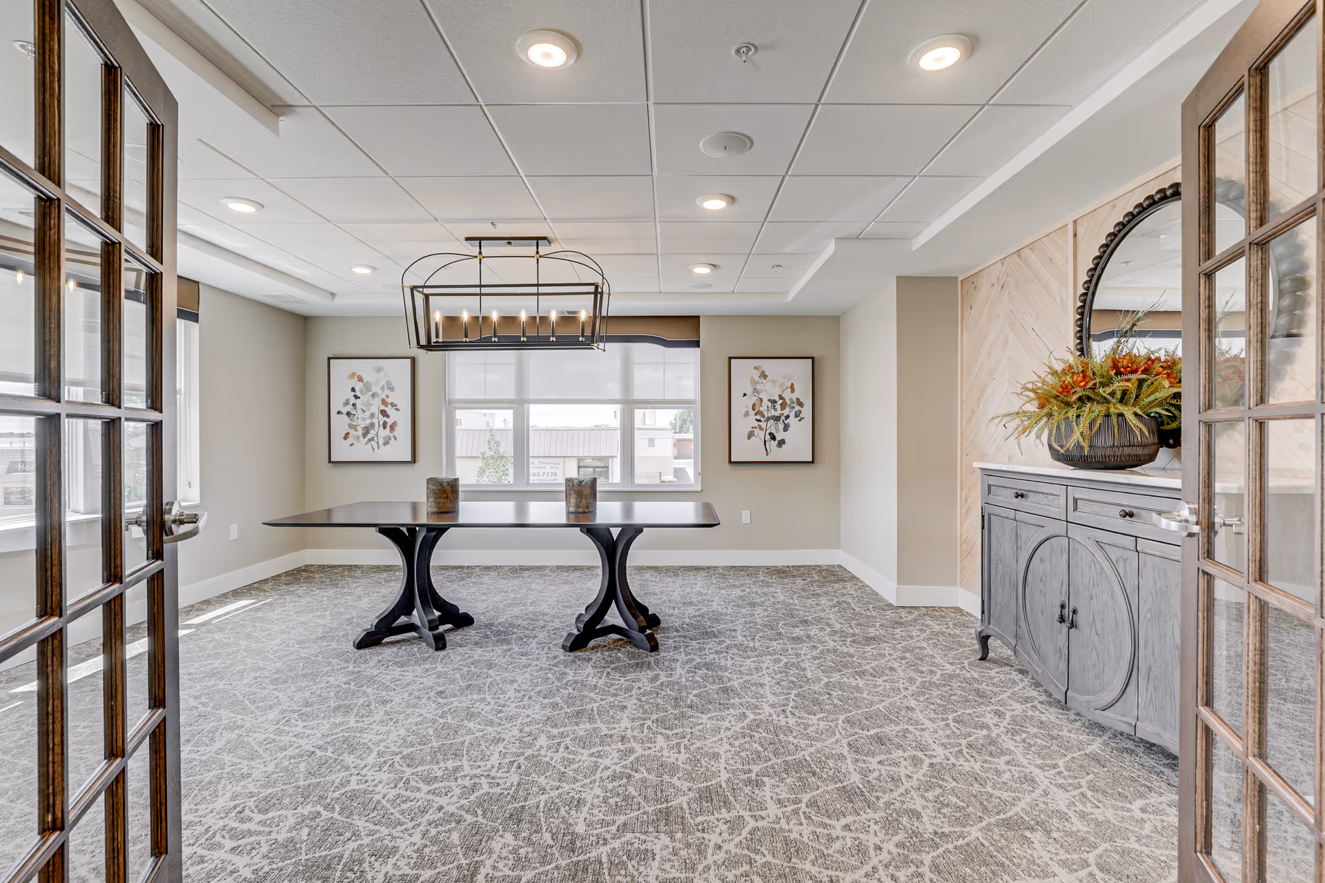 A bright, modern interior room with a large rectangular table in the center supported by two black pedestal bases. The room has patterned carpet flooring, beige walls, and a white ceiling with recessed lighting. Two framed botanical prints hang on the far wall between two large windows with white blinds. To the right, there is a gray sideboard with a large round mirror above it and a decorative plant arrangement. The room is viewed through partially open wooden framed glass double doors.