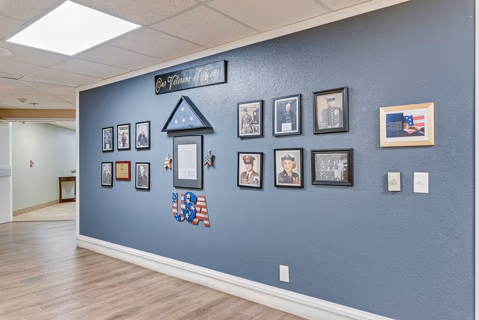 Interior hallway wall display dedicated to veterans, featuring framed photographs, a folded American flag in a triangular case, patriotic decorations including stars and USA letters, and a sign that reads 'Our Veterans of Honor'. The wall is painted blue and the floor has light wood laminate.