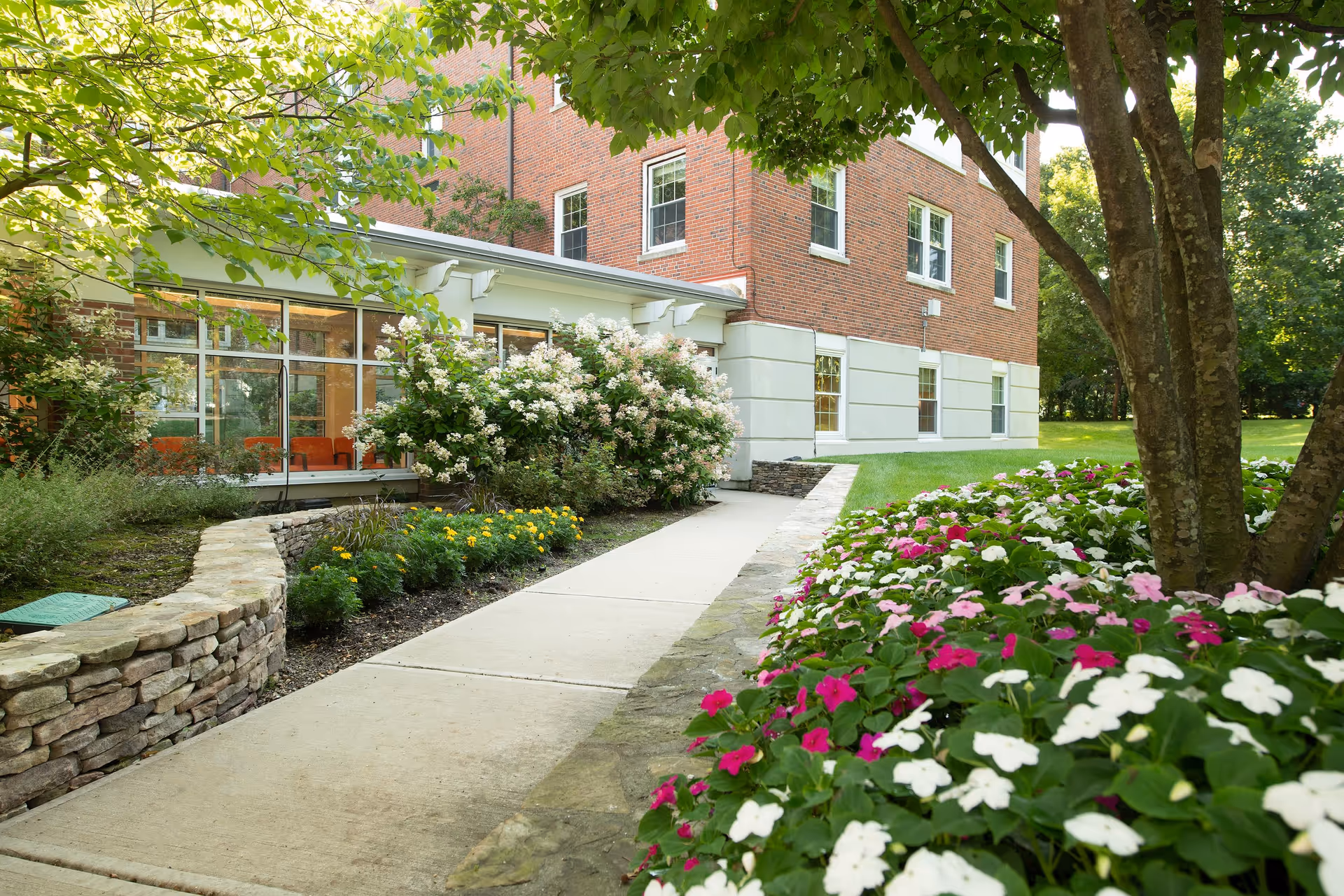 A paved walkway leading to a brick building with large windows, surrounded by well-maintained flower beds with pink, white, and yellow flowers, and green trees providing shade.