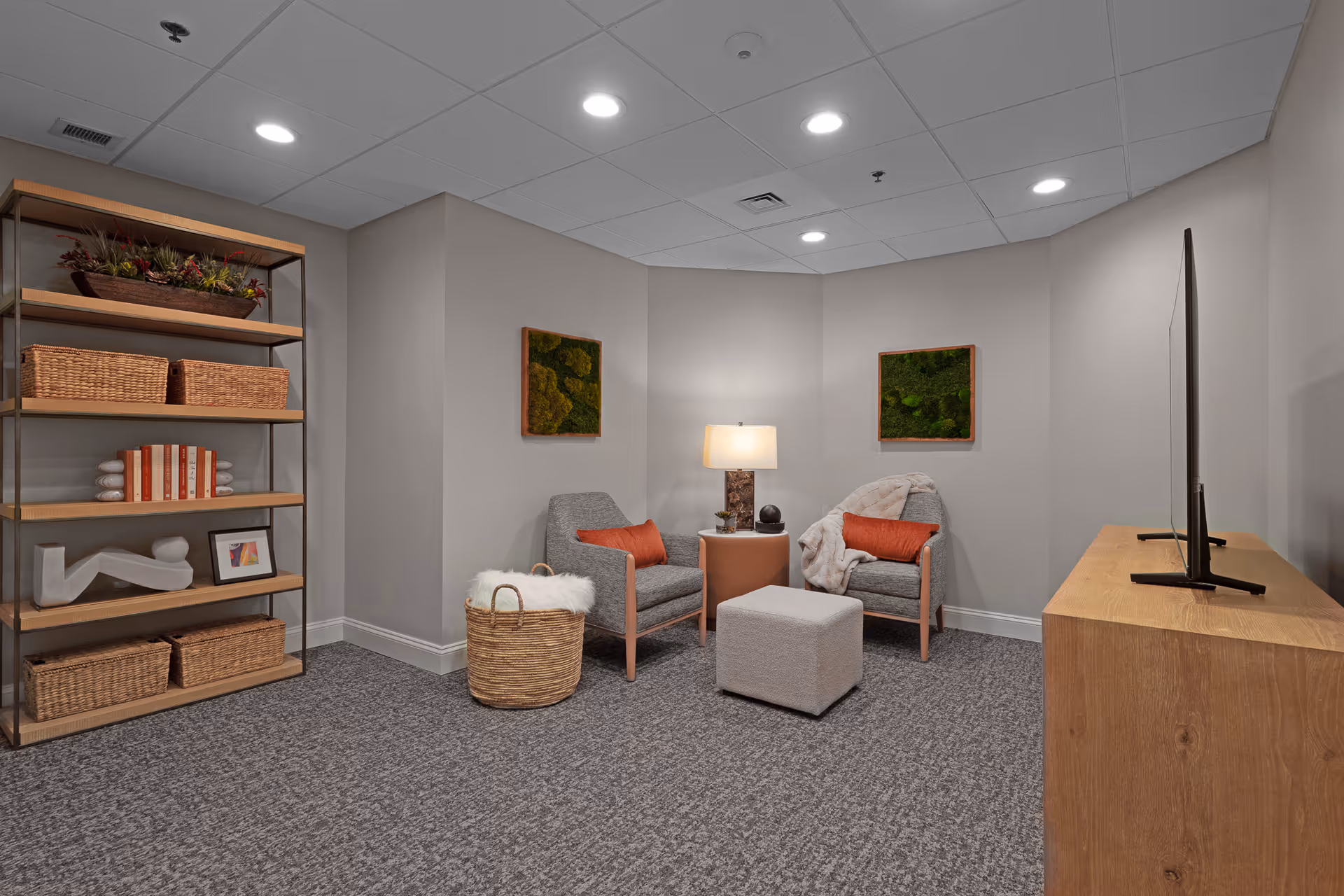 A cozy sitting area with two gray armchairs each with an orange pillow, a small round wooden side table with a lamp, a gray ottoman, and a wooden TV stand with a flat-screen TV. On the left side, there is a wooden shelving unit with baskets, books, and decorative items. The walls are light gray with two green moss art pieces hanging. The floor is carpeted in gray.
