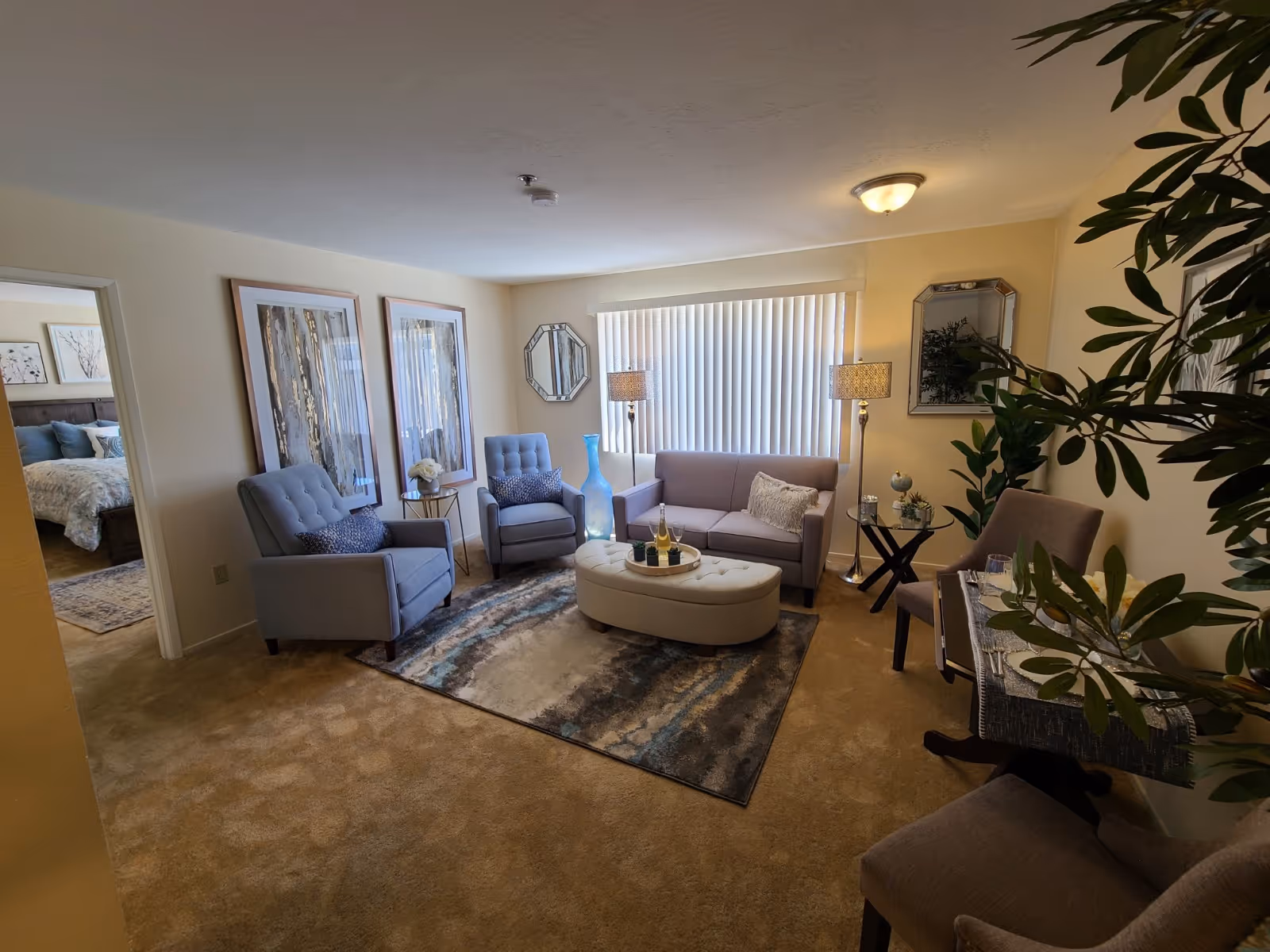 Well-lit furnished living room with sofa, armchairs, ottoman on a rug, lamps, artwork, and plants.