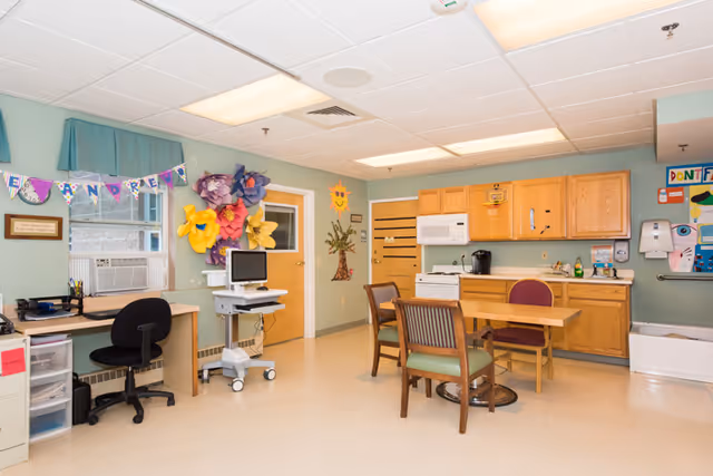 A bright and clean room with light green walls and a tiled ceiling. The room features a wooden table with three chairs, a desk with a black office chair, a computer on a mobile stand, and wooden cabinets with a microwave and coffee maker. Colorful paper flowers and a banner decorate the wall near a door with a window. There are also various posters and a hand sanitizer dispenser on the walls.