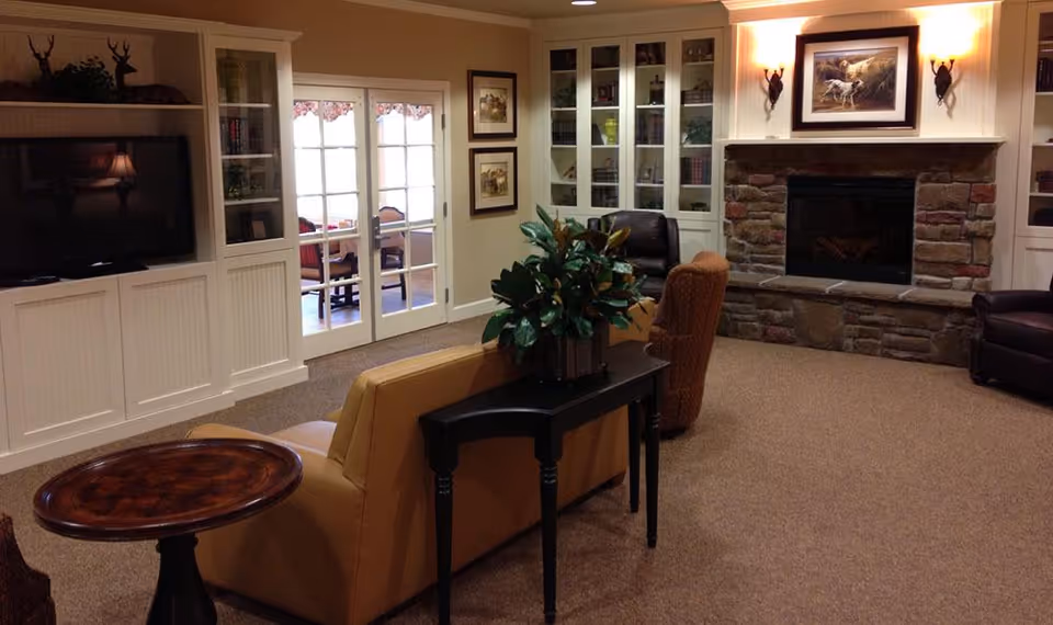 A cozy living room with a stone fireplace, a painting of dogs above the mantel, built-in white cabinets with glass doors, a flat-screen TV, and comfortable seating including a tan sofa and armchairs. There is a small black table with a plant on it behind the sofa and a round wooden side table. French doors lead to another room with a table and chairs.