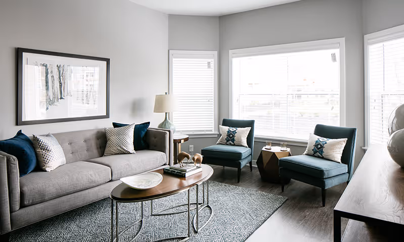 Bright living room with a gray sofa, two teal armchairs, a wooden coffee table, area rug, and large windows.