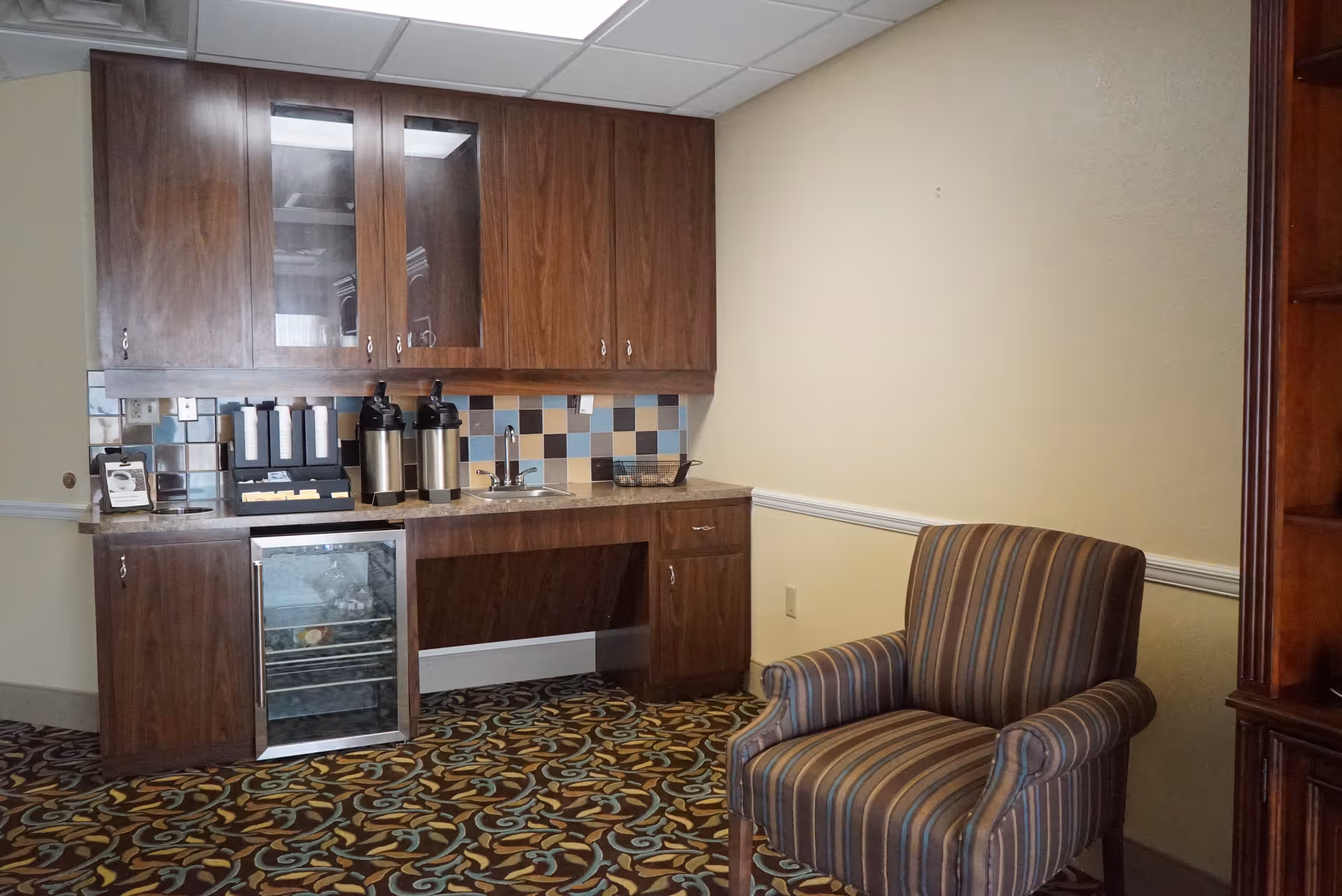 Small kitchenette with wood cabinets, a mini-fridge, coffee dispensers and a sink next to a striped armchair in a carpeted lounge area.