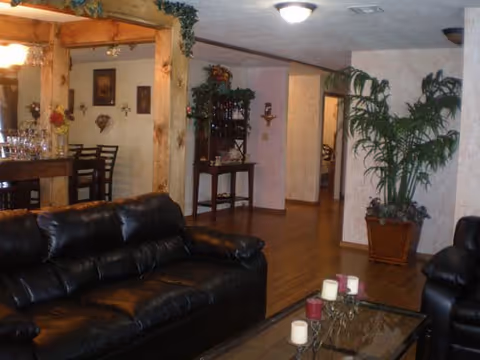 Interior living room with black leather sofas, a glass coffee table with candles, and a view into a dining/bar area and hallway with potted plants.