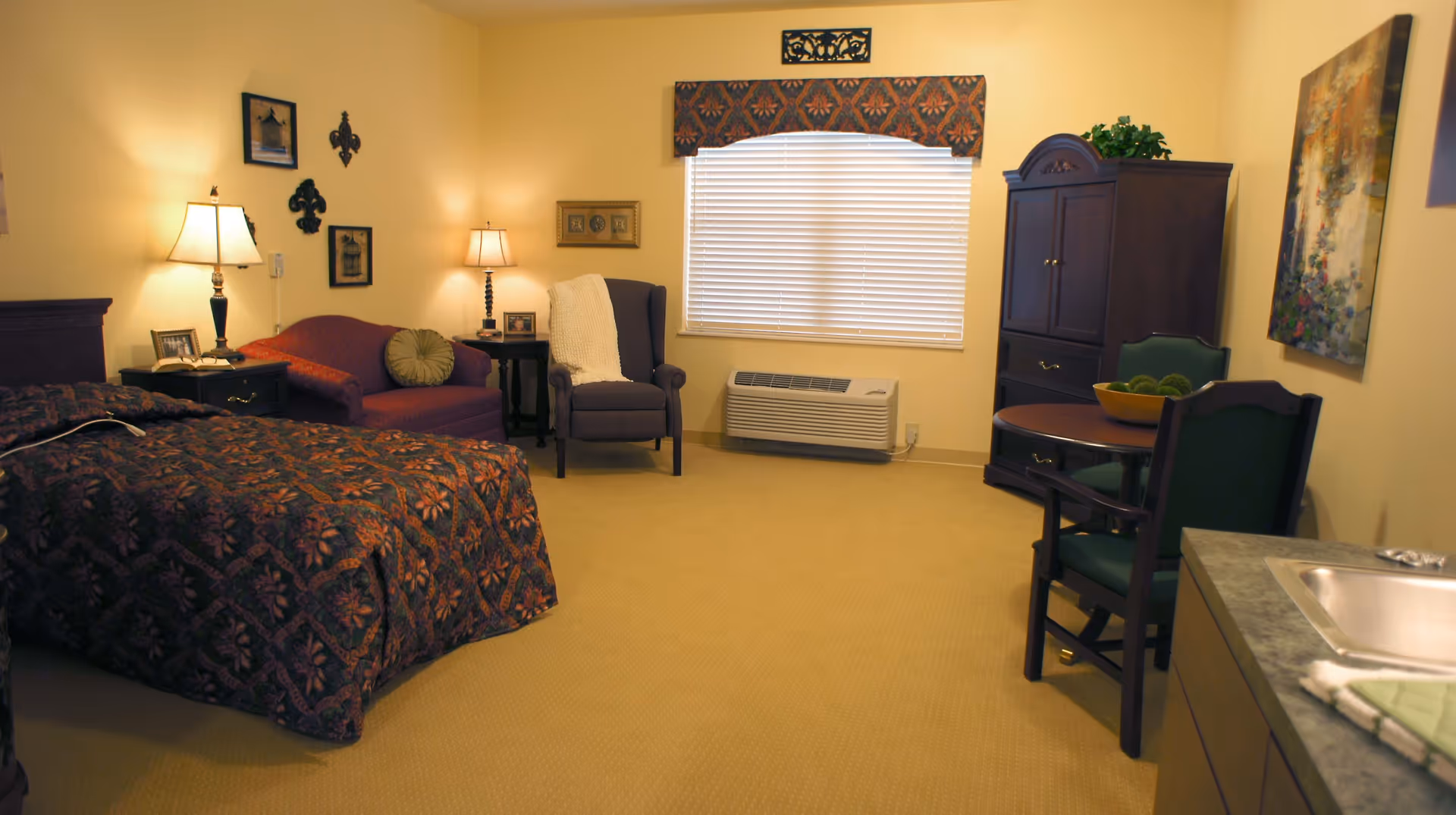 A cozy bedroom in a senior living facility featuring a bed with a patterned bedspread, a small sofa with a round pillow, a wingback chair with a white throw, two lamps on side tables, a window with blinds and a decorative valance, a wooden cabinet, a round table with two chairs, and a small kitchenette area with a sink.