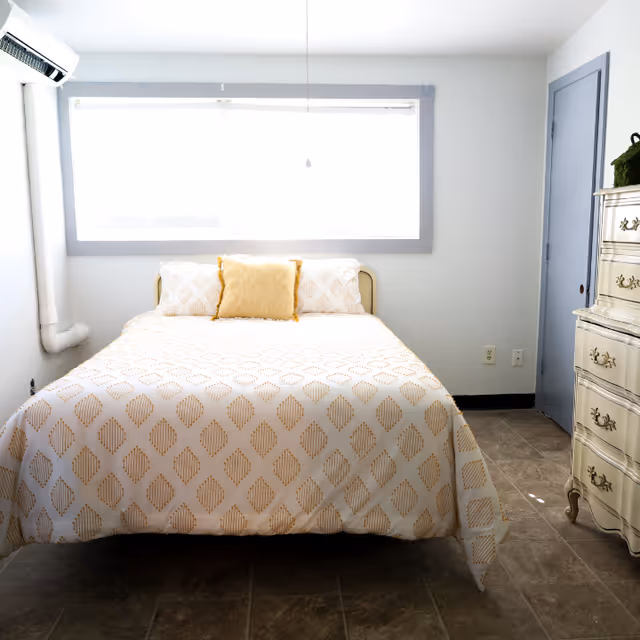 A bright bedroom with a bed covered in a white and gold patterned bedspread, two matching pillows, and a yellow accent pillow. There is a large window above the bed letting in natural light, a white air conditioning unit on the left wall, and a vintage-style cream-colored dresser on the right side of the room. The floor is tiled, and the walls are painted light gray.