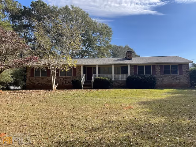 Single-story brick house with a front porch, surrounded by trees and a large grassy yard under a partly cloudy blue sky.