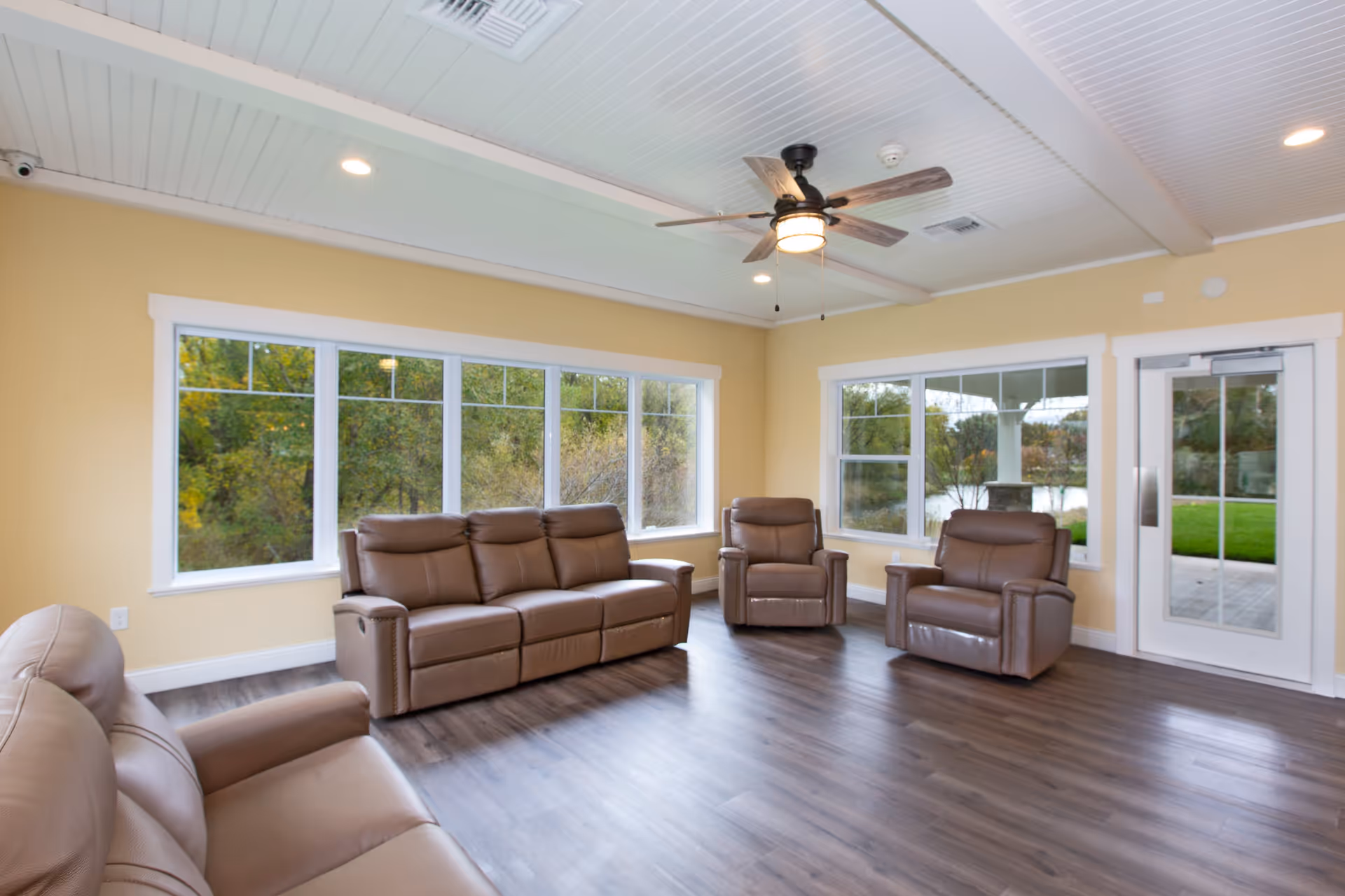 A bright living room with large windows showing green trees outside. The room has light yellow walls, a white ceiling with recessed lights and a ceiling fan with wooden blades. There are three brown leather recliners and a matching brown leather sofa arranged on a dark wood floor. A glass door leads outside to a patio area with green grass.