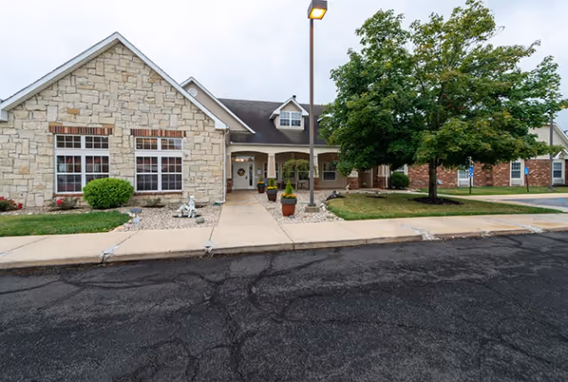 Front entrance of a single-story senior living building with a stone facade, covered walkway, potted plants, and a tree in the lawn.