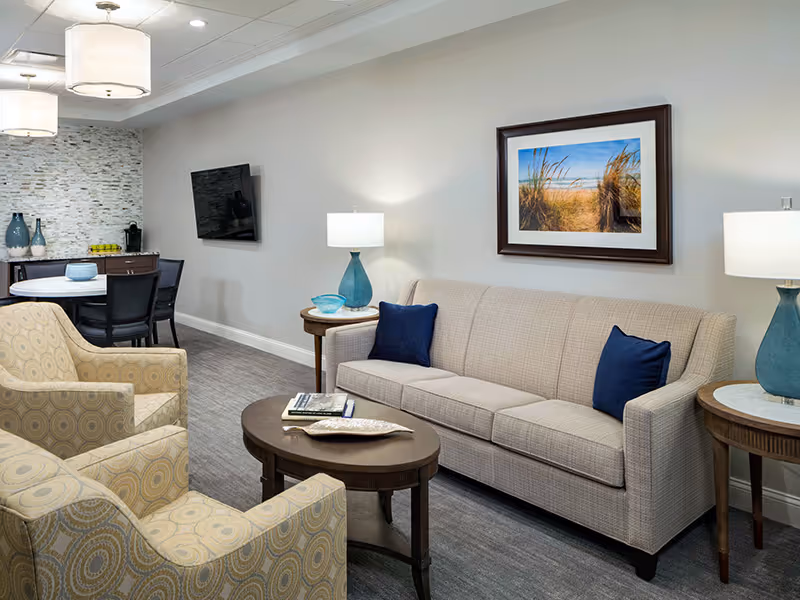 A cozy living room area featuring a beige sofa with two blue pillows, two patterned armchairs, a wooden coffee table with books and a decorative item, two side tables with blue lamps, a wall-mounted flat screen TV, a round dining table with chairs, and a framed picture of a beach scene on the wall.