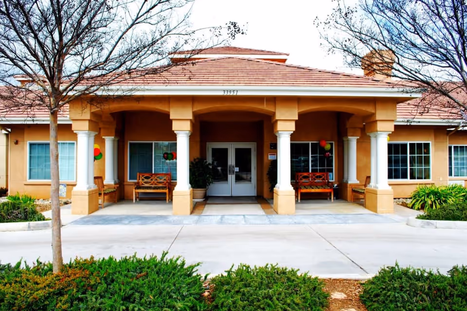 Front exterior view of Wildwood Canyon Villa Assisted Living and Memory Care building with a covered entrance supported by columns, benches on the porch, leafless trees, and shrubs in the foreground.