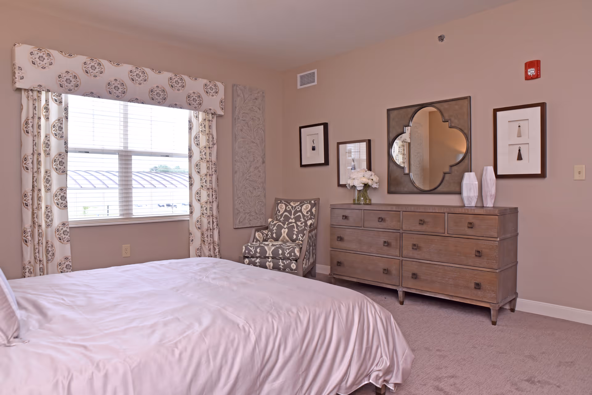 A bedroom with a bed covered in white bedding, a window with patterned curtains, a patterned armchair in the corner, and a wooden dresser with a decorative mirror, framed pictures, and vases on top.