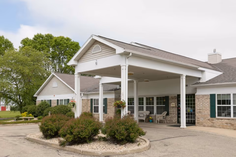 Covered drop-off entrance (porte-cochere) of a single-story brick senior living building with shrubs, hanging planters, and outdoor seating.
