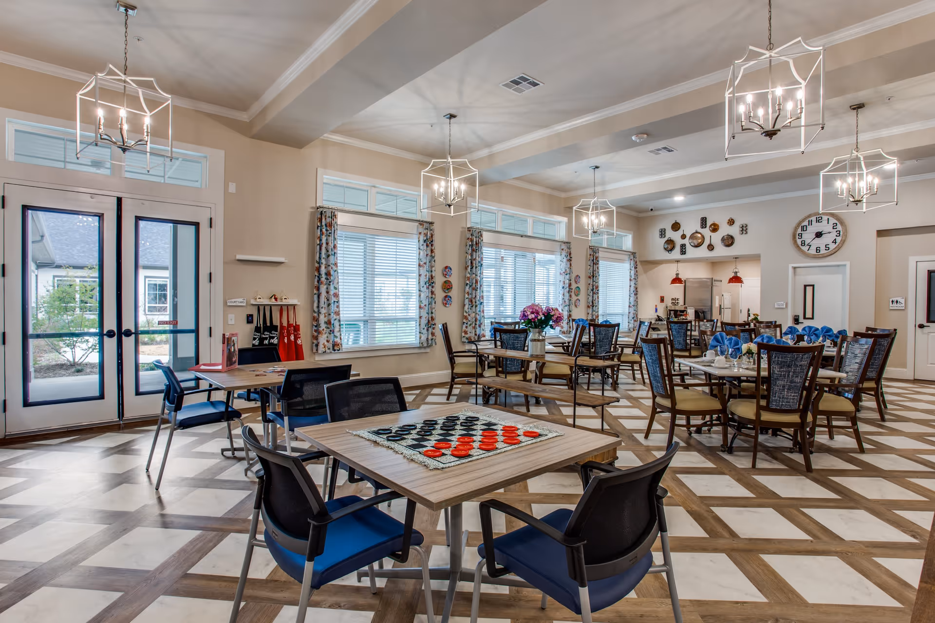 A spacious and well-lit common area in a senior living facility with multiple tables and chairs arranged for dining and social activities. The room features large windows with floral curtains, modern chandeliers, a checkers game set on one table, and a kitchen area in the background. The floor has a checkered pattern, and there is a large clock on the wall near the kitchen and restroom doors.