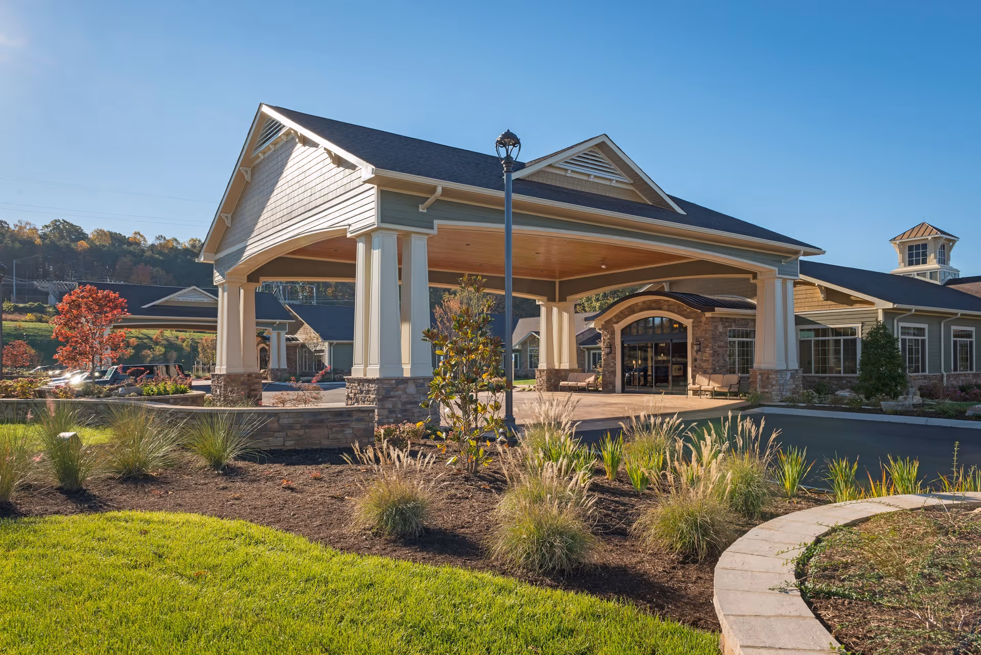 Exterior view of a senior living facility entrance with a covered drop-off area supported by columns. The building has a stone and siding facade with large windows and a landscaped garden with grass, shrubs, and small trees in the foreground under a clear blue sky.