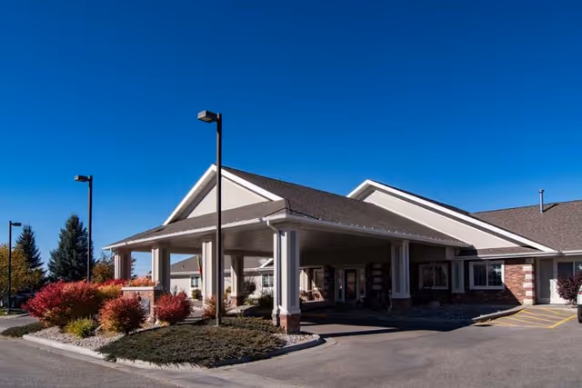 Front entrance and porte-cochere of a single-story senior living building with landscaped shrubs under a clear blue sky.